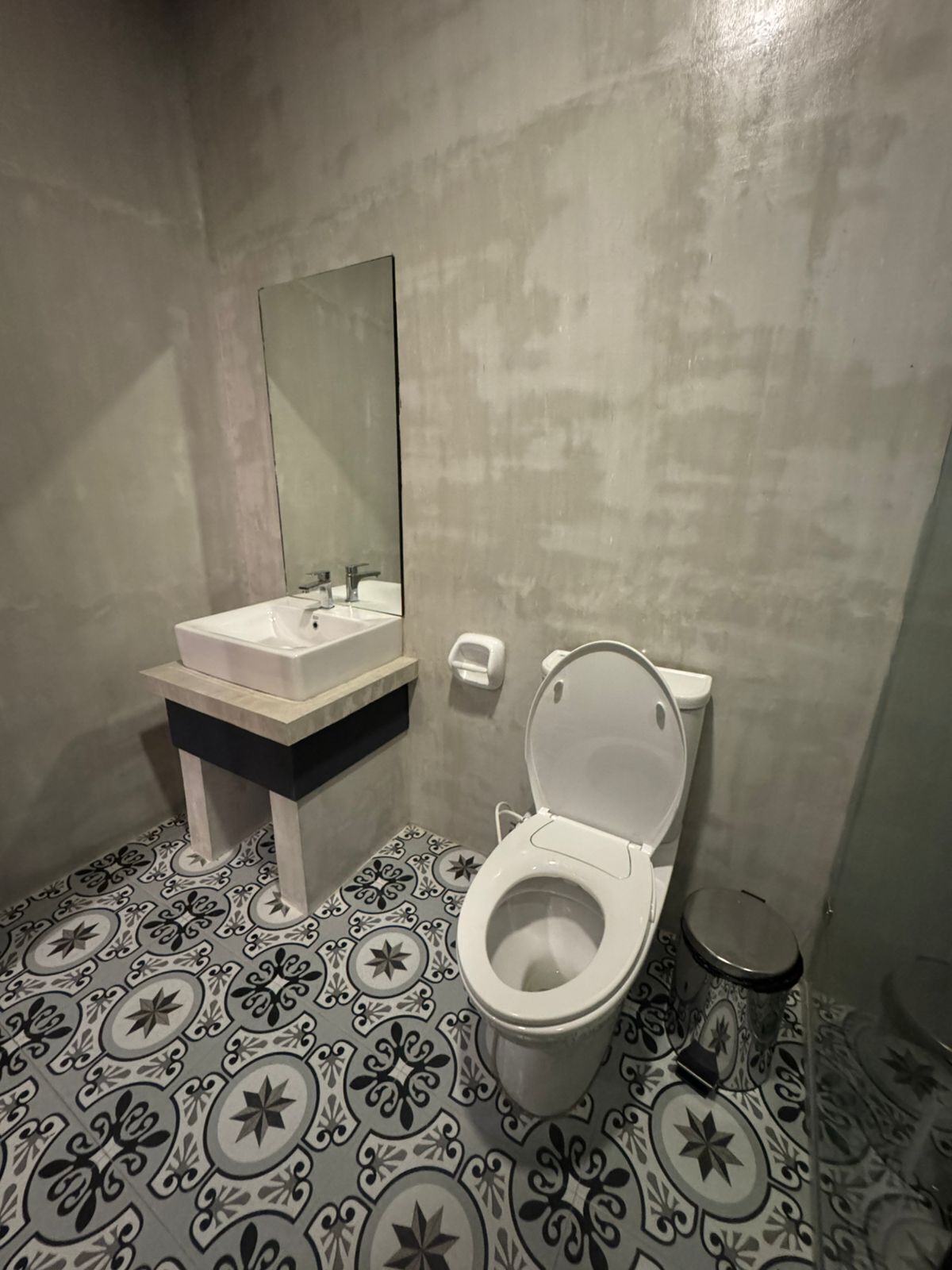Ein Badezimmer mit Toilette, Waschbecken und Spiegel. Der Boden ist mit einem gemusterten Fliesenbelag versehen.