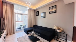 A modern living room with a black sofa, a glass coffee table, and a view of a cityscape through large windows.