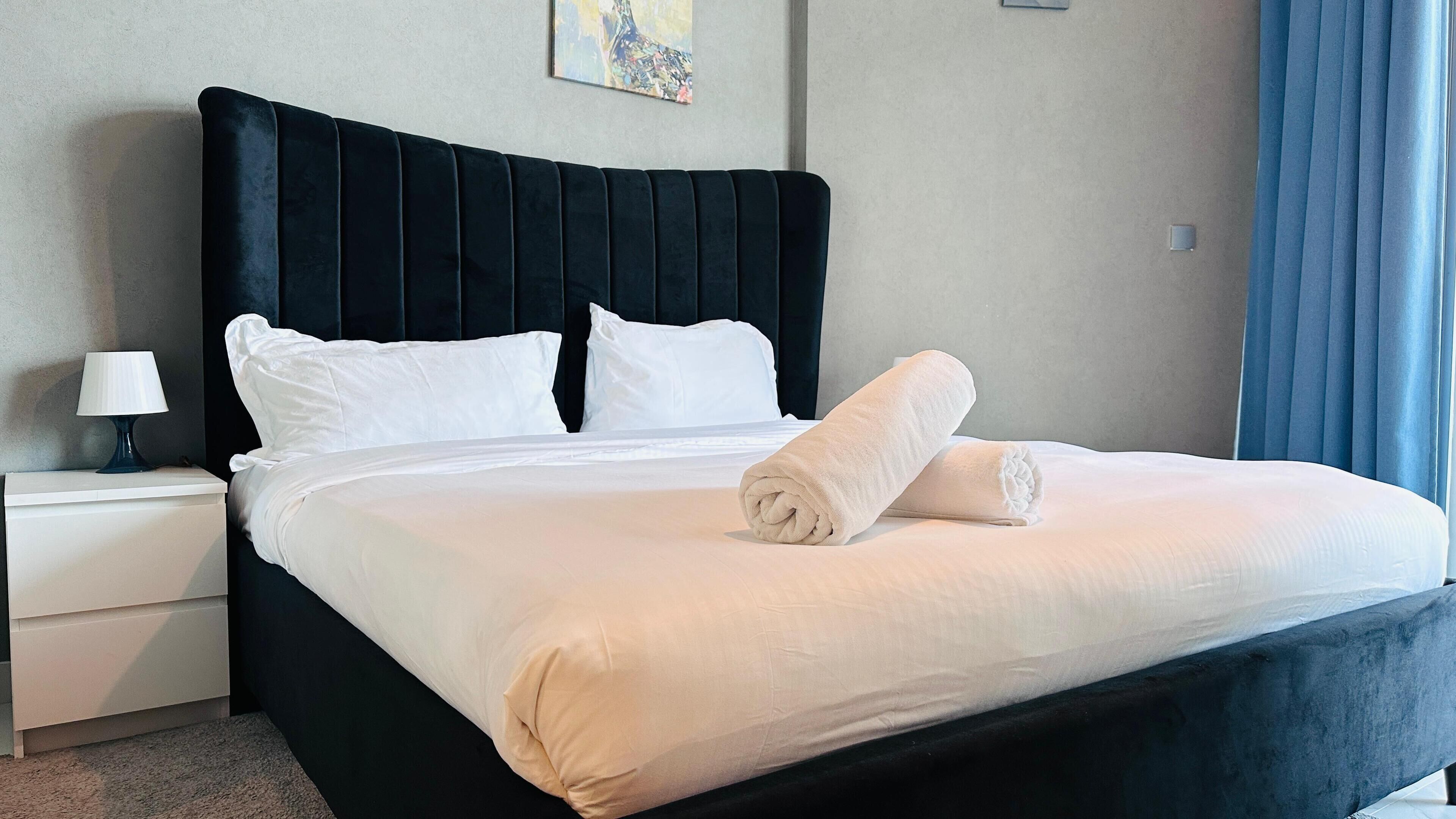 A neatly made bed with white linens and two rolled towels, a dark upholstered headboard, a bedside table with a lamp, and a blue curtain.