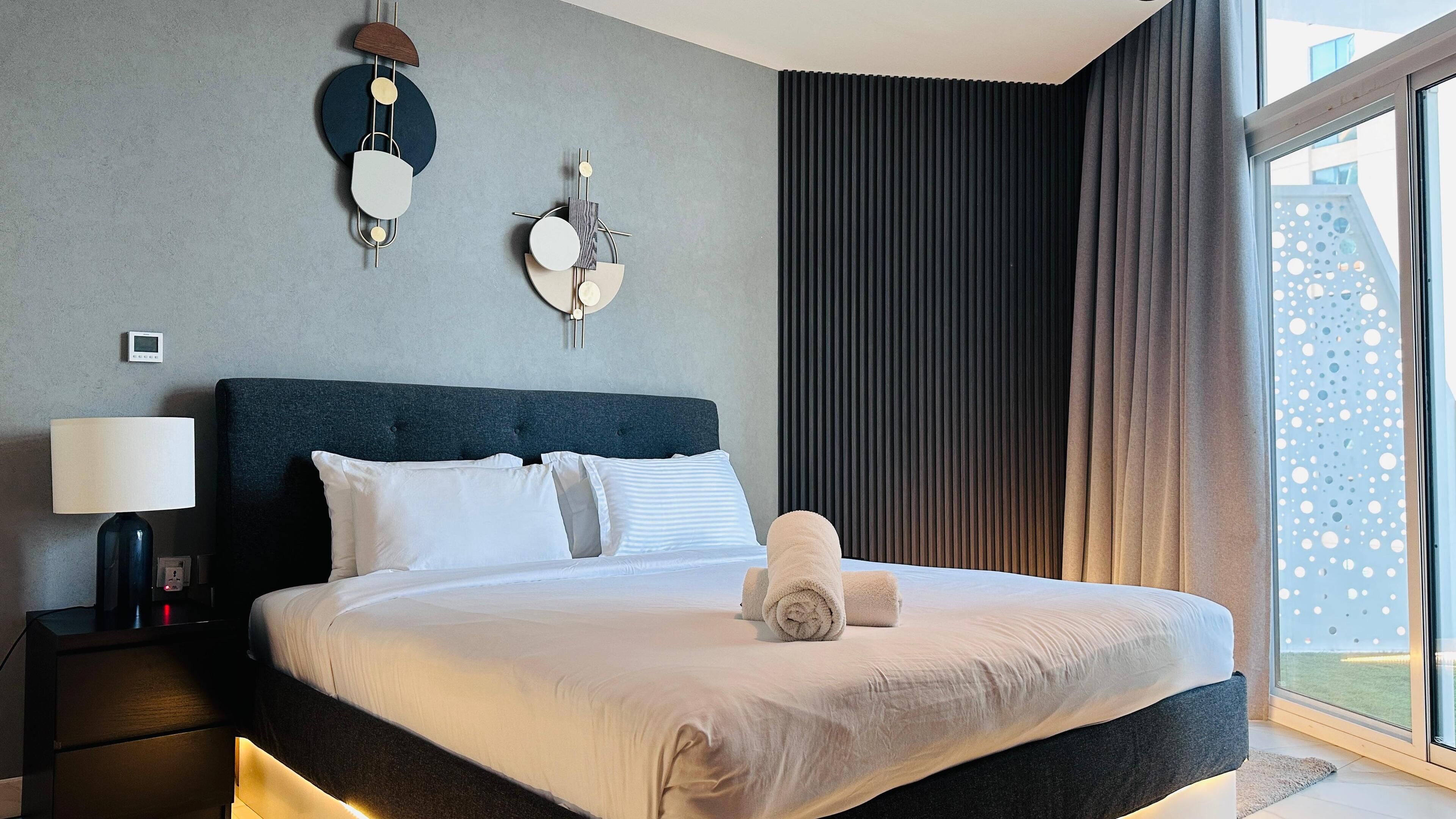 A modern hotel room with a large bed, a bedside table with a lamp, and a view of the city through the window.