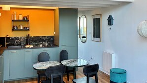 A modern kitchen with a built-in island, a dining area with a round table and chairs, and a loft space with a hanging light fixture. - 3* Apartment for 2–4 people, 25 m² terrace—50 m from the beach and the GR34 trail. Dogs Welcome (Carantec)