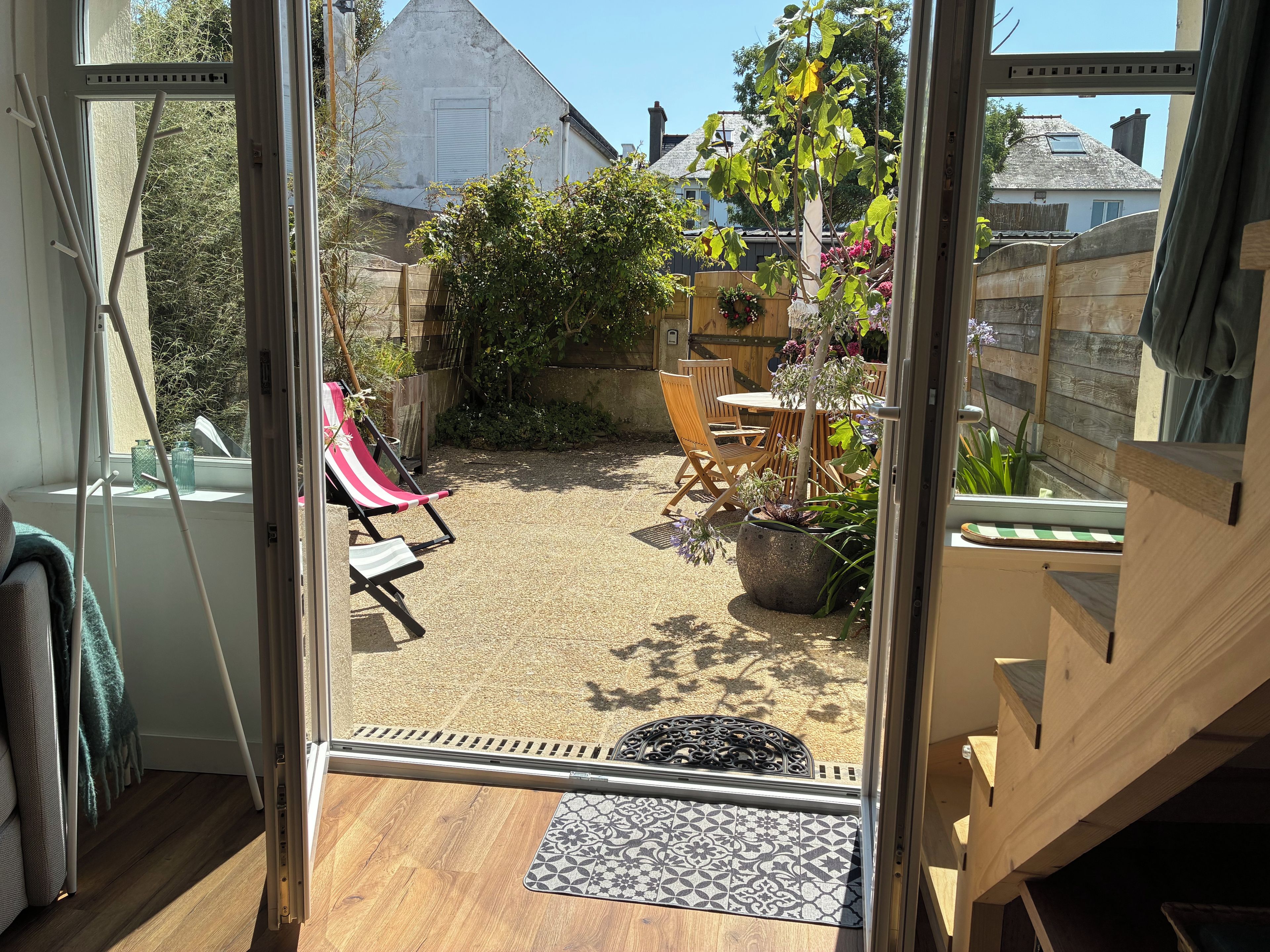 A view through a glass door into a backyard with a patio area, a wooden table, and a chair.