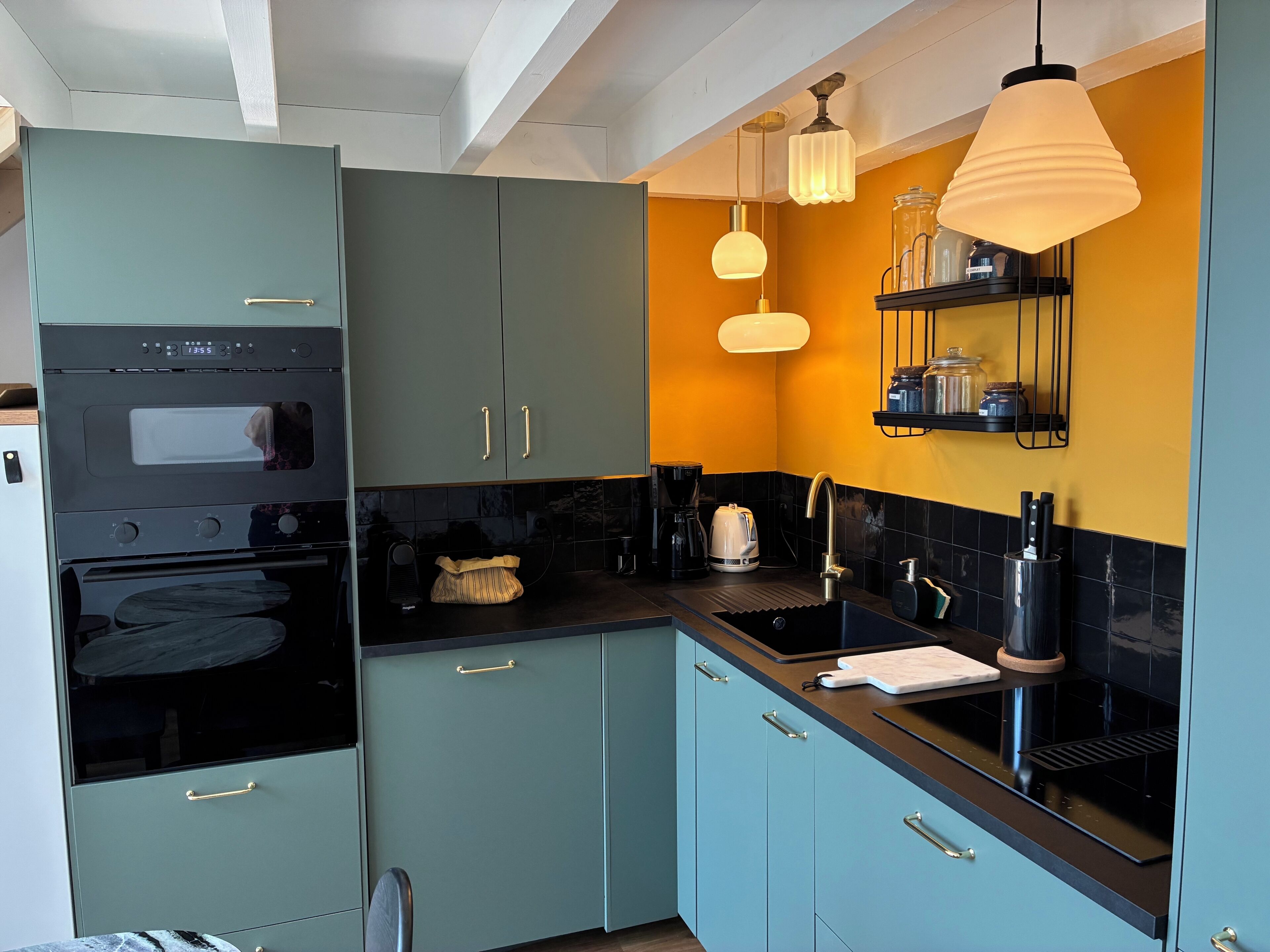 A modern kitchen with teal cabinets, black countertops, a built-in oven, a coffee maker, and pendant lighting.