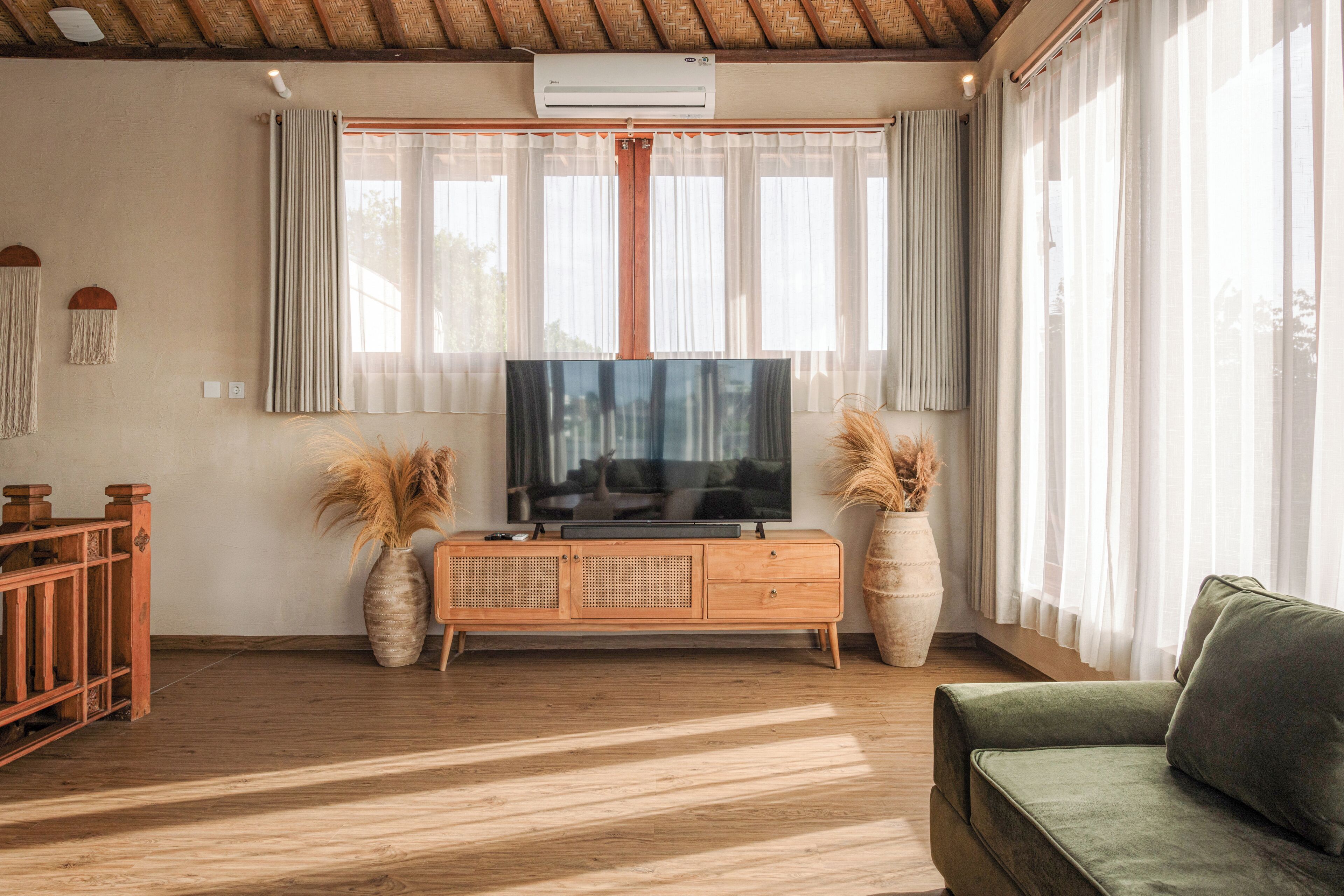 A living room with a wooden TV stand, a green sofa, and two large windows with sheer curtains.