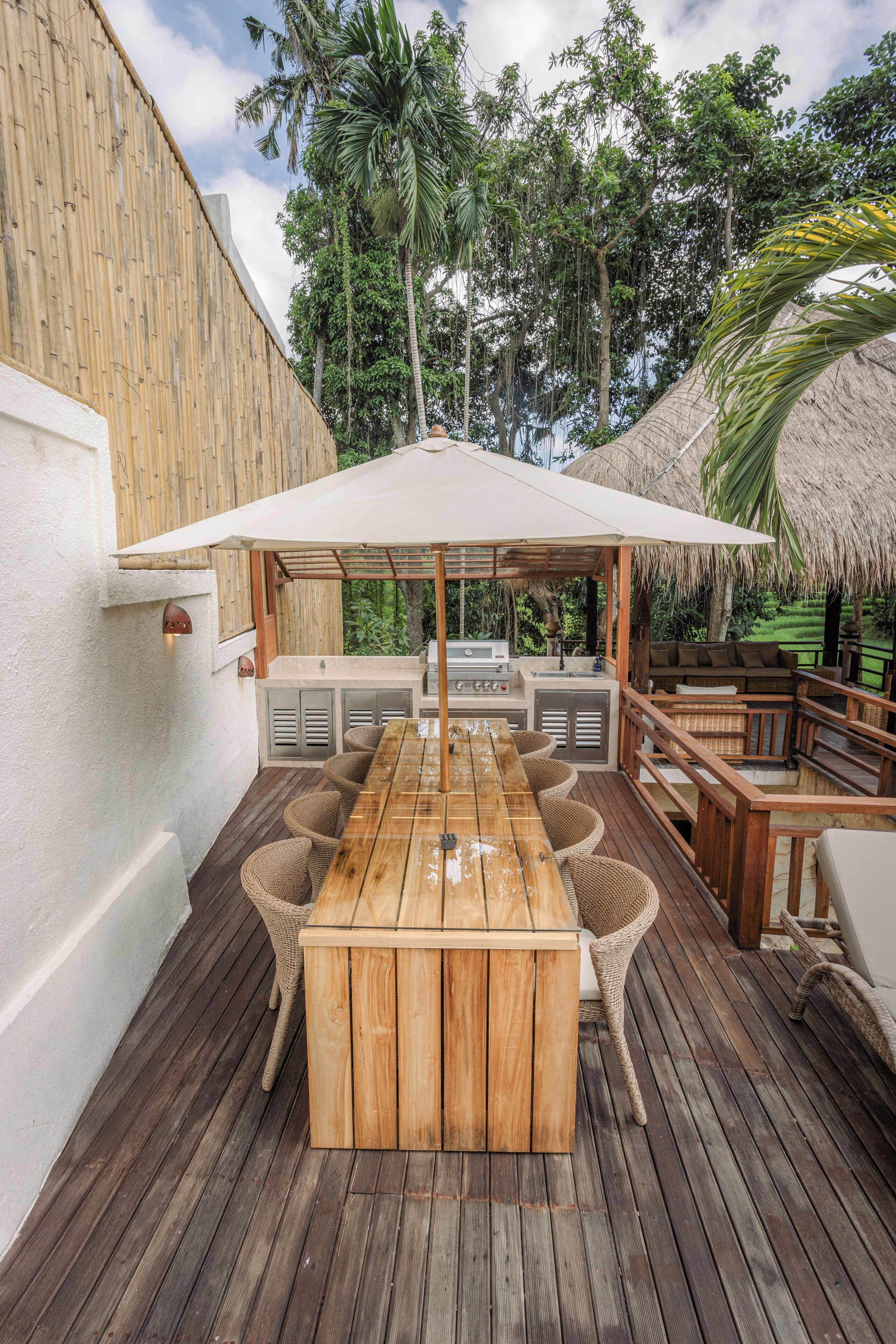 Una terraza de madera con una mesa, sillas, una parrilla y una estructura de techo de paja.