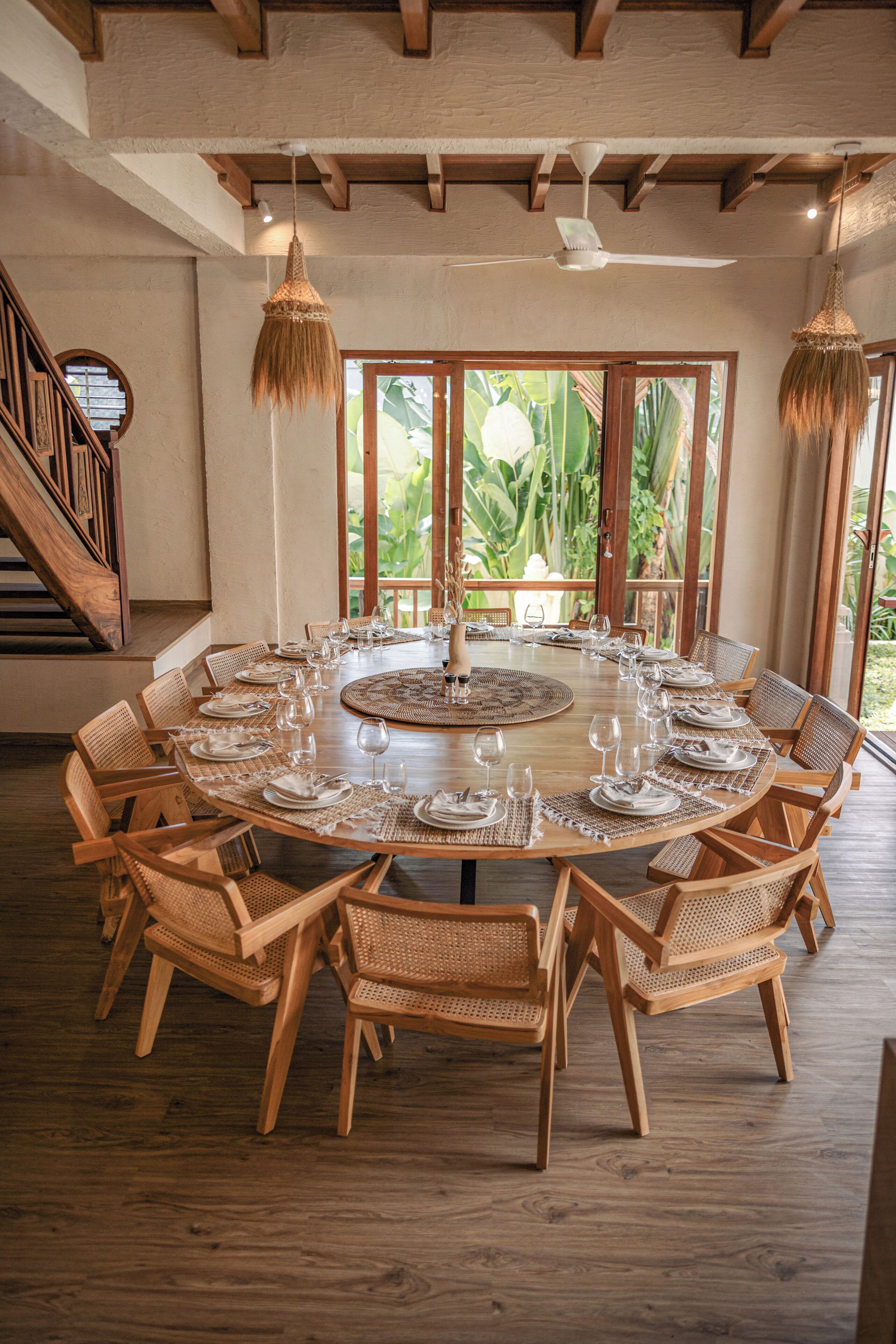 A round wooden dining table set for six with woven placemats, surrounded by six wicker chairs.