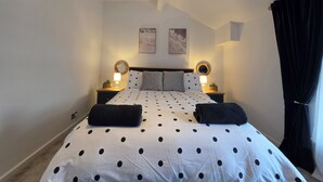 Un dormitorio con cama, dos mesitas de noche, un velador, un espejo montado en la pared y una ventana con cortinas.