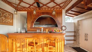 Una barra de madera con tres taburetes, una cocina al fondo y un espejo grande sobre la barra.