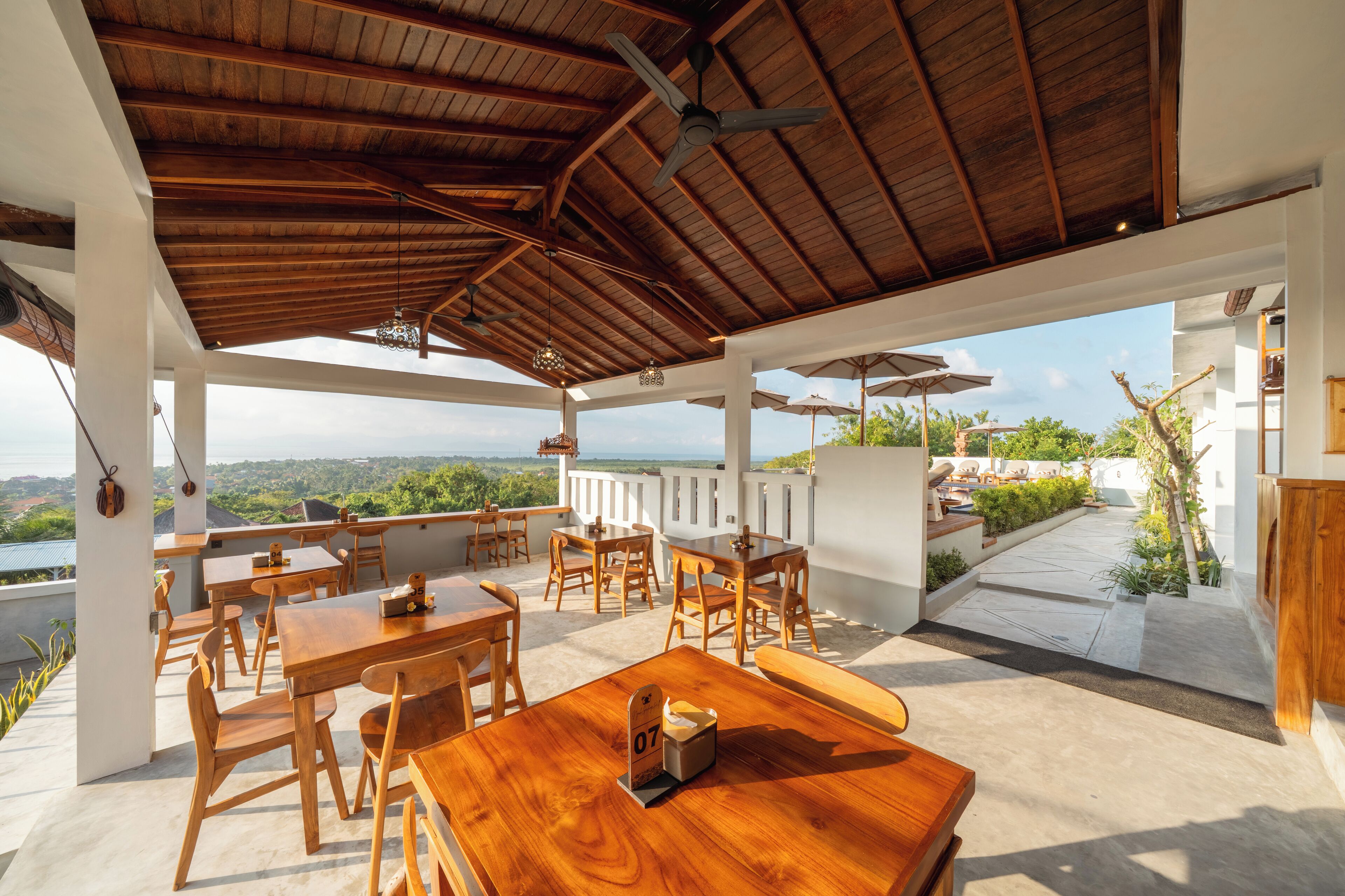 An outdoor patio with wooden tables and chairs, a ceiling fan, and a view of the landscape.