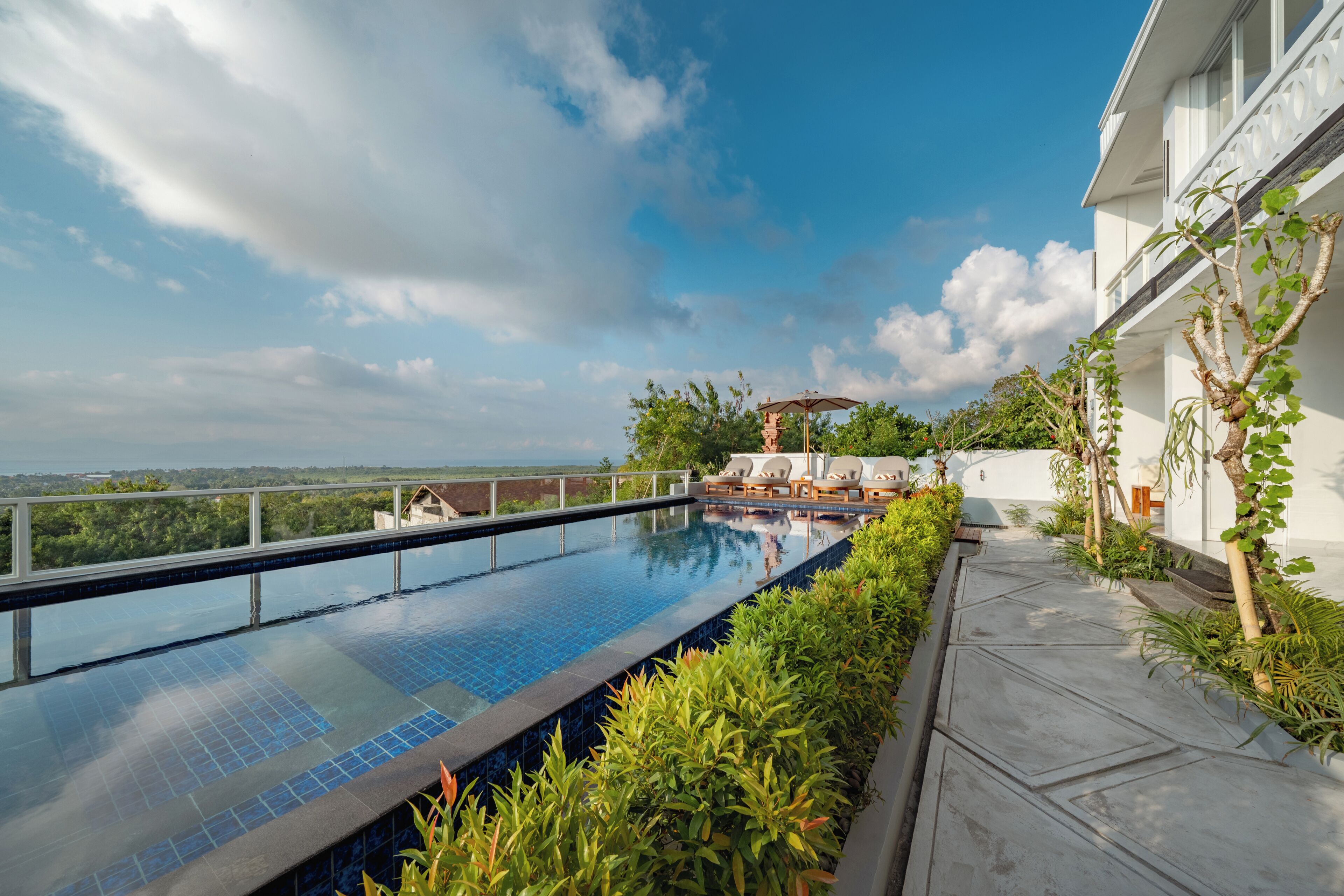 A rooftop infinity pool with a view of the ocean, surrounded by lush greenery and a white building.