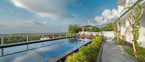 A rooftop infinity pool with a view of the ocean, surrounded by lush greenery and a white building.