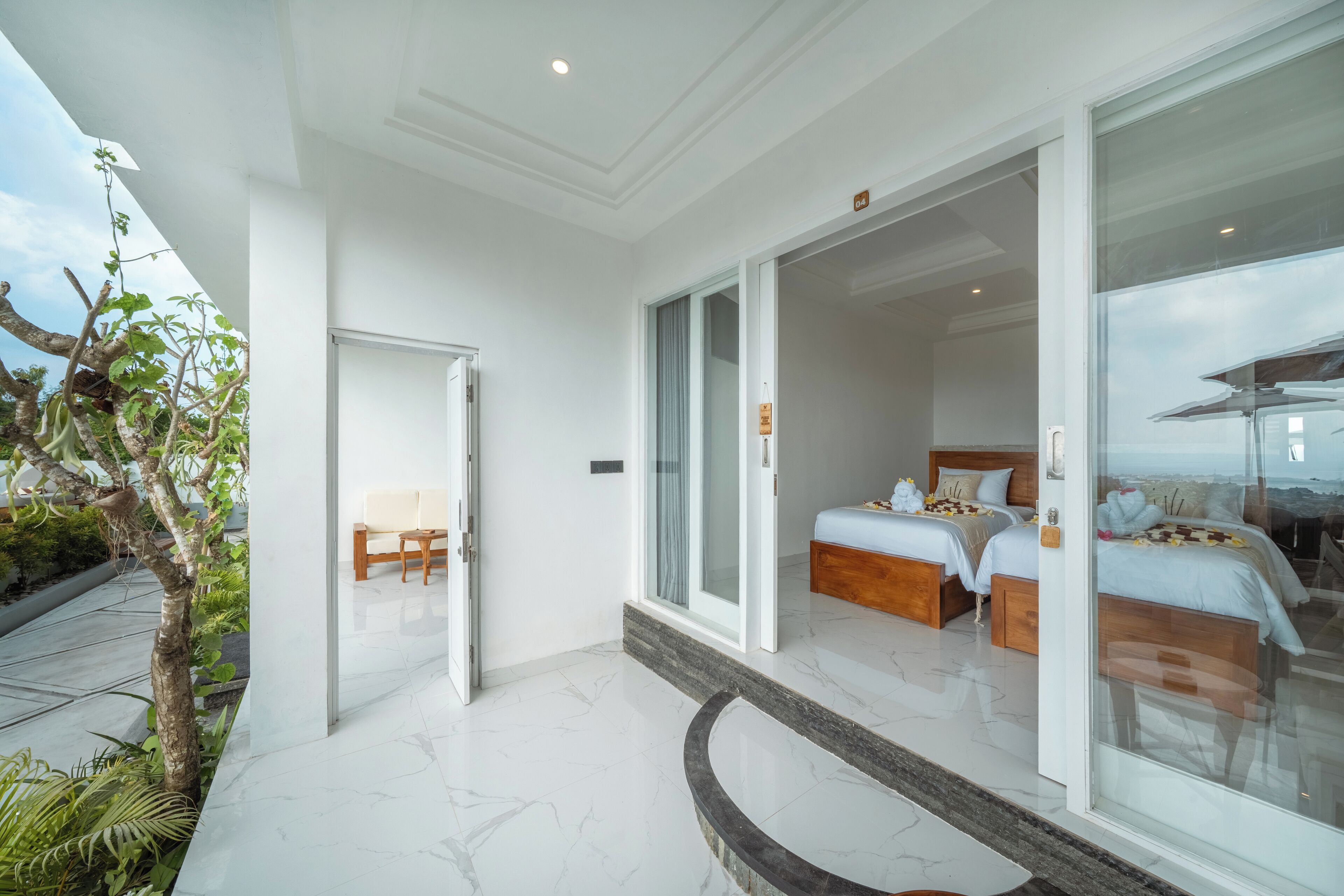 A modern hotel room with a large sliding glass door leading to a balcony, a separate sitting area, and a spacious bedroom with two beds.