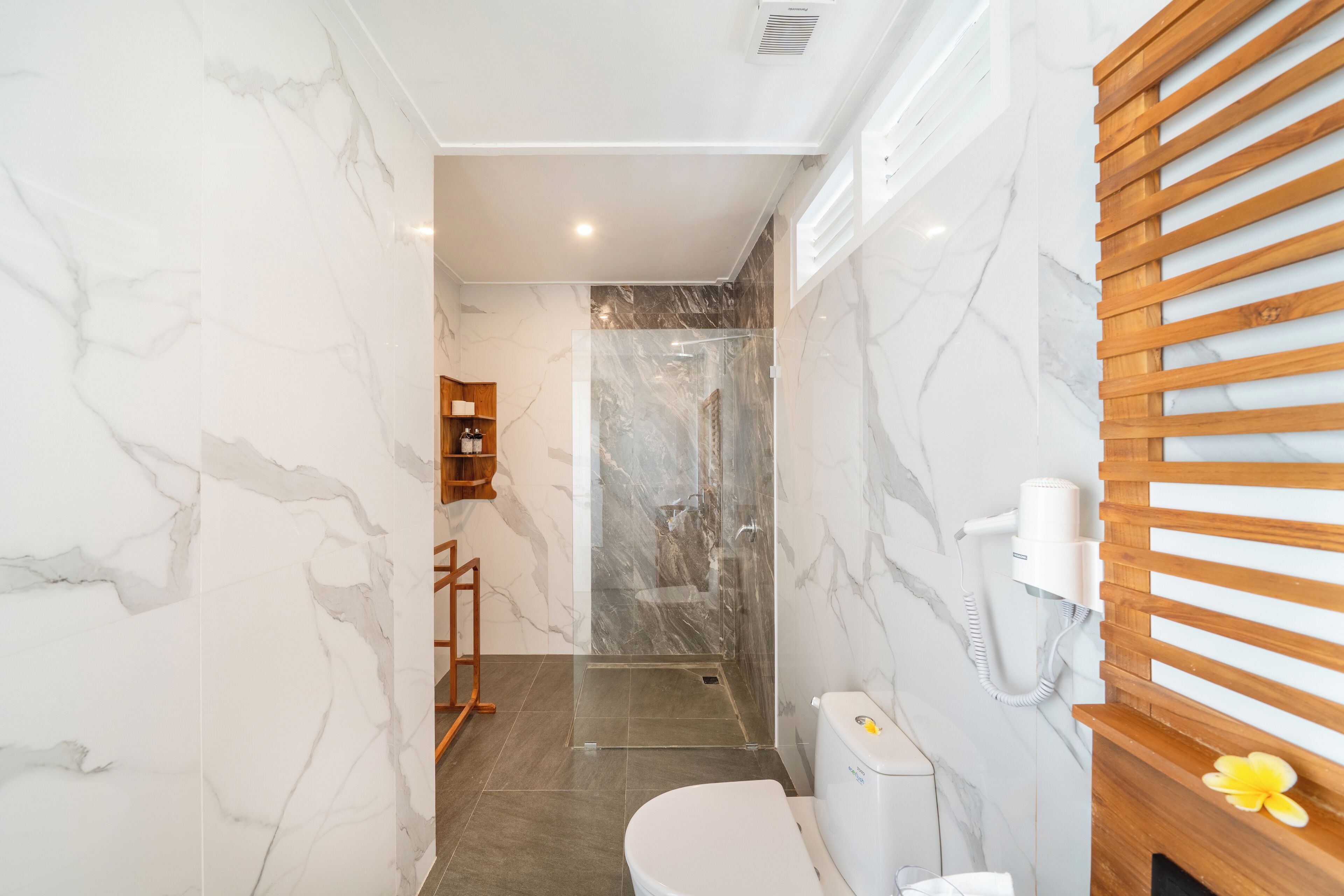 Un baño moderno con paredes de mármol, cabina de ducha de cristal, tocador de madera y un inodoro con secador de pelo montado en la pared.