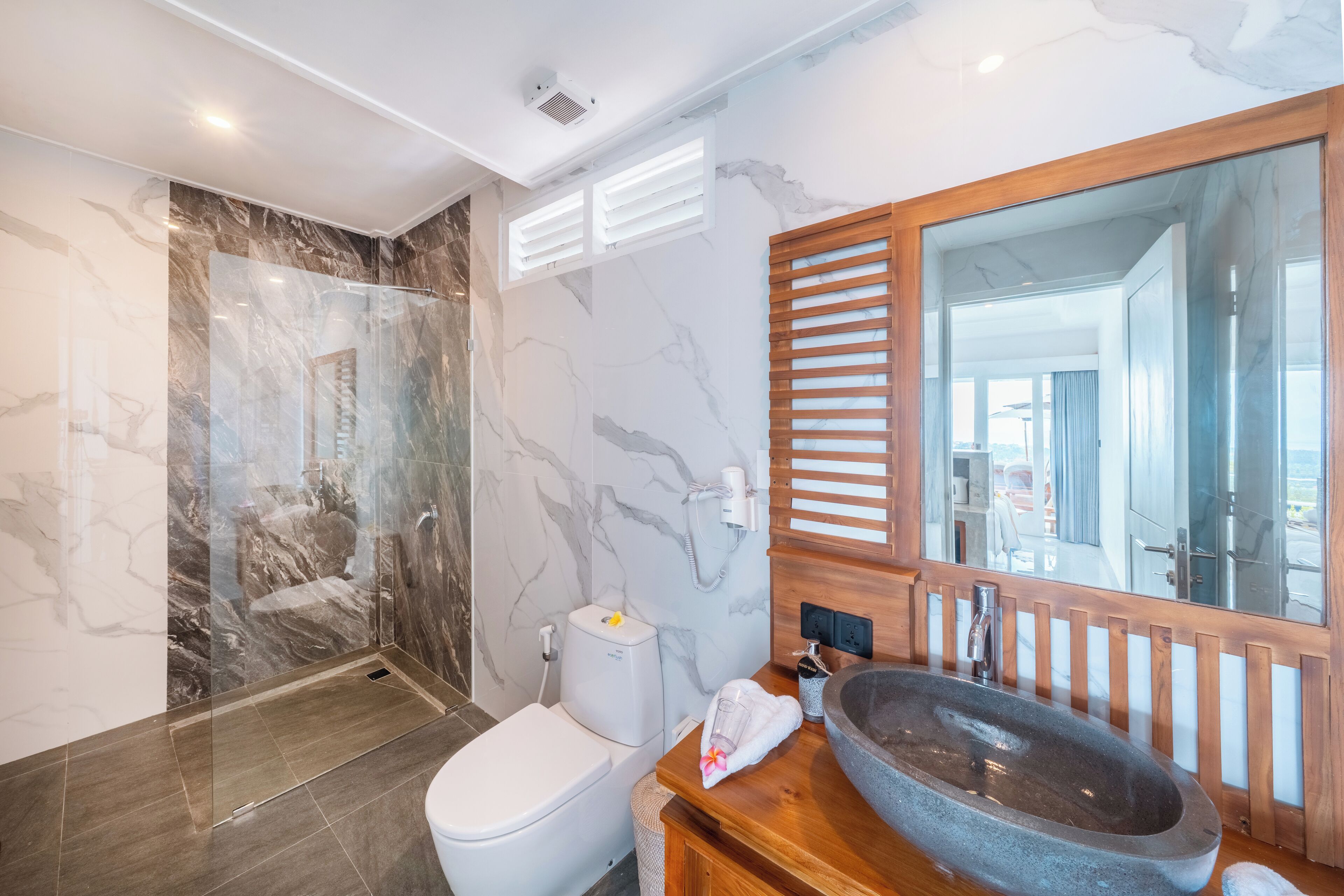 Un baño moderno con cabina de ducha de vidrio, pared con azulejos de mármol, tocador de madera con un lavabo grande y un espejo amplio.