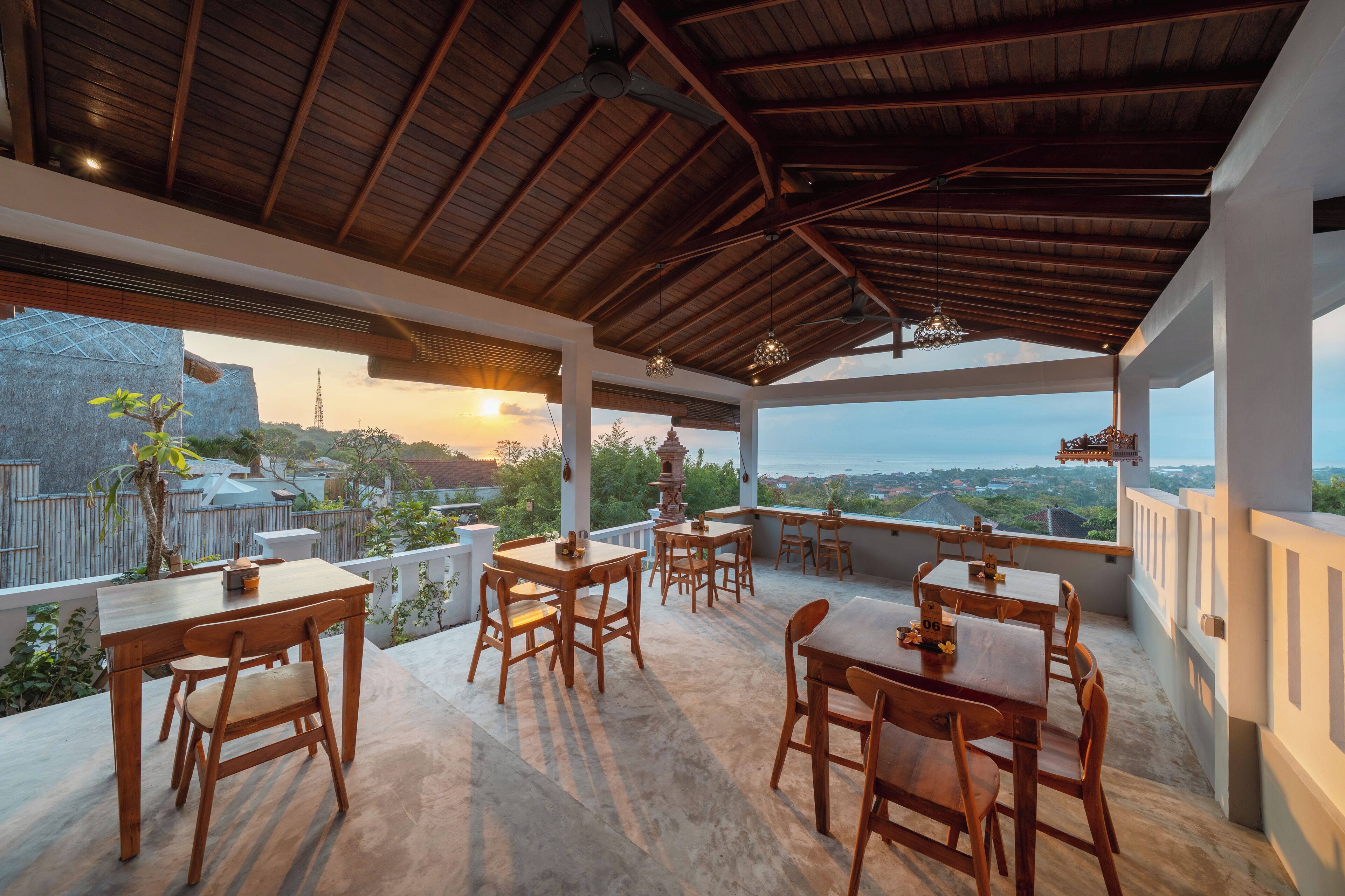 A covered outdoor dining area with wooden tables and chairs, large windows offering a view of the landscape, and a wooden ceiling structure.