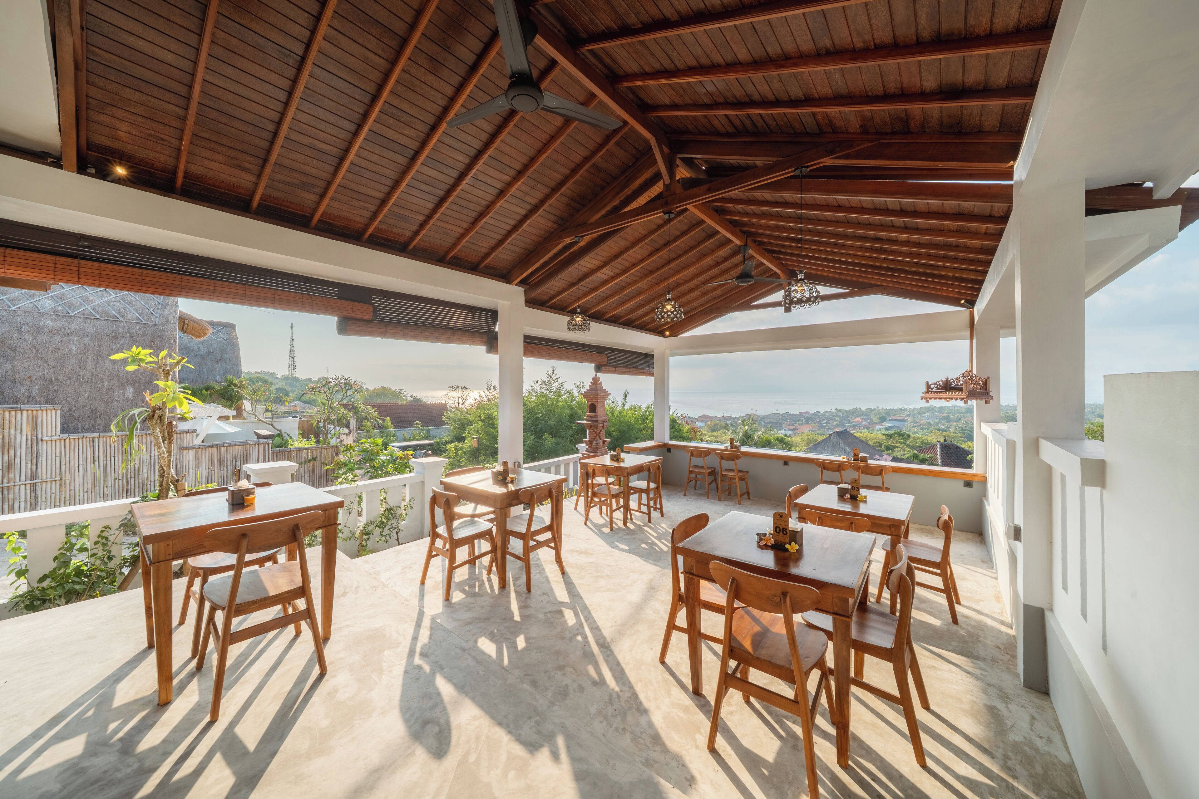 Un espacio de comedor al aire libre cubierto, con mesas y sillas de madera, y ventanas amplias que ofrecen vistas del paisaje circundante.