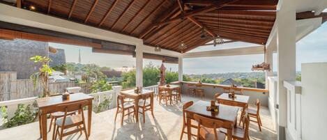 Un espacio de comedor al aire libre cubierto, con mesas y sillas de madera, y ventanas amplias que ofrecen vistas del paisaje circundante.