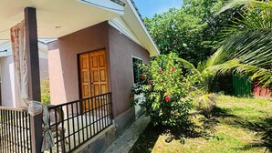 Una casa con puerta de madera, un balcón con barandilla metálica y un jardín con diversas plantas y una palmera.