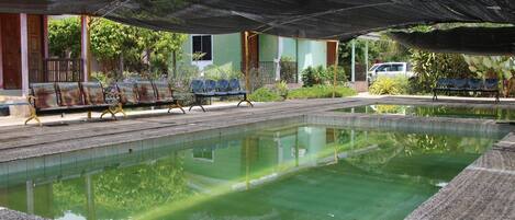 Zona de piscina con un área de estar cubierta y un edificio al fondo.