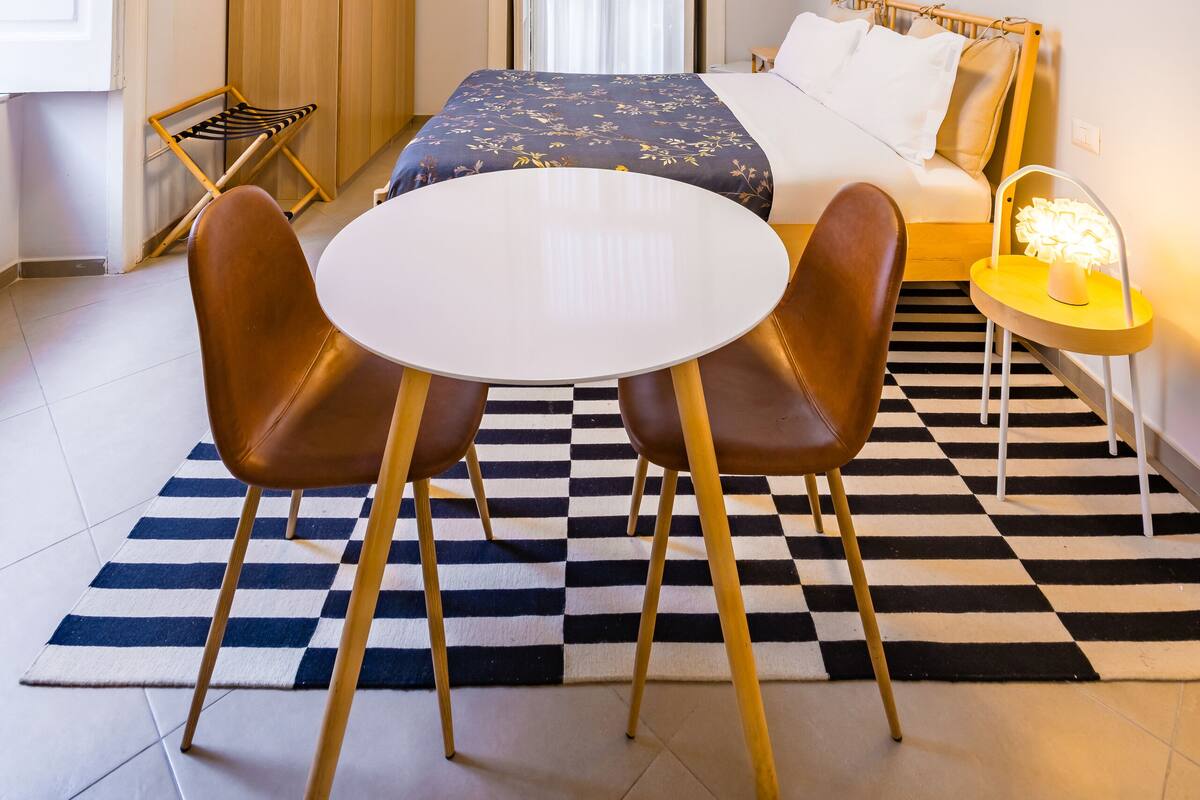 A compact hotel room with a bed, a small round table with two chairs, a wardrobe, and a checkered floor.