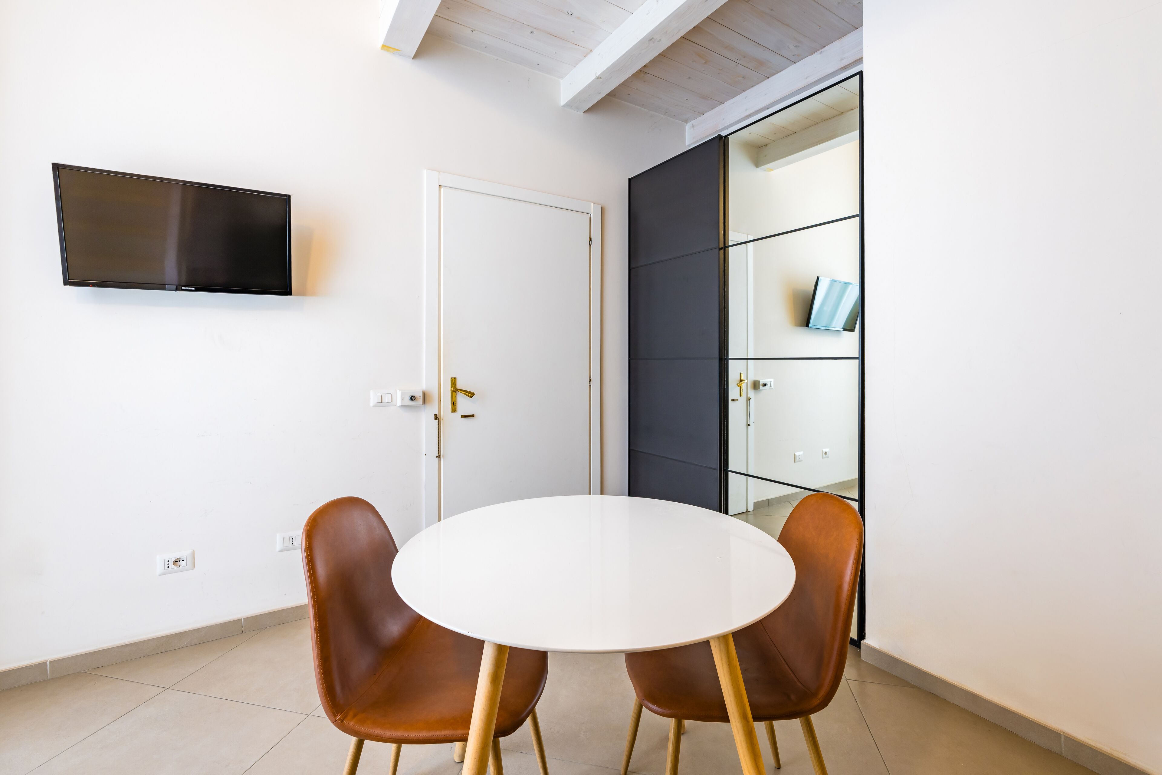 A room with a flat-screen TV mounted on the wall, a white door, a glass cabinet with a blue object, and two brown chairs around a white table.