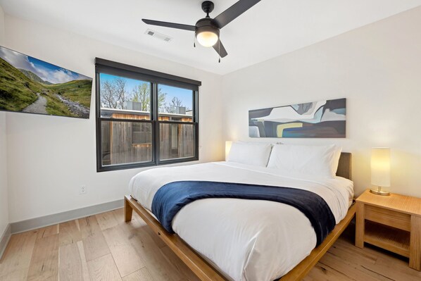 A bedroom with a bed, a ceiling fan, a window with a view, and a bedside table with a lamp.
