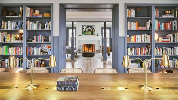 Una habitación amplia con una gran mesa de madera, dos sillas y una estantería llena de libros. Se ve una chimenea al fondo, y la habitación tiene un diseño moderno.