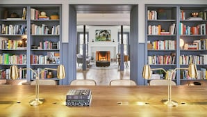 Una habitación amplia con una gran mesa de madera, dos sillas y una estantería llena de libros. Se ve una chimenea al fondo, y la habitación tiene un diseño moderno.