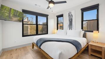 A bedroom with a bed, a ceiling fan, two windows, a nightstand, and a wall-mounted TV.