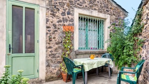 A stone building with a green door, a window with green bars, a table with a green tablecloth, and two green chairs.