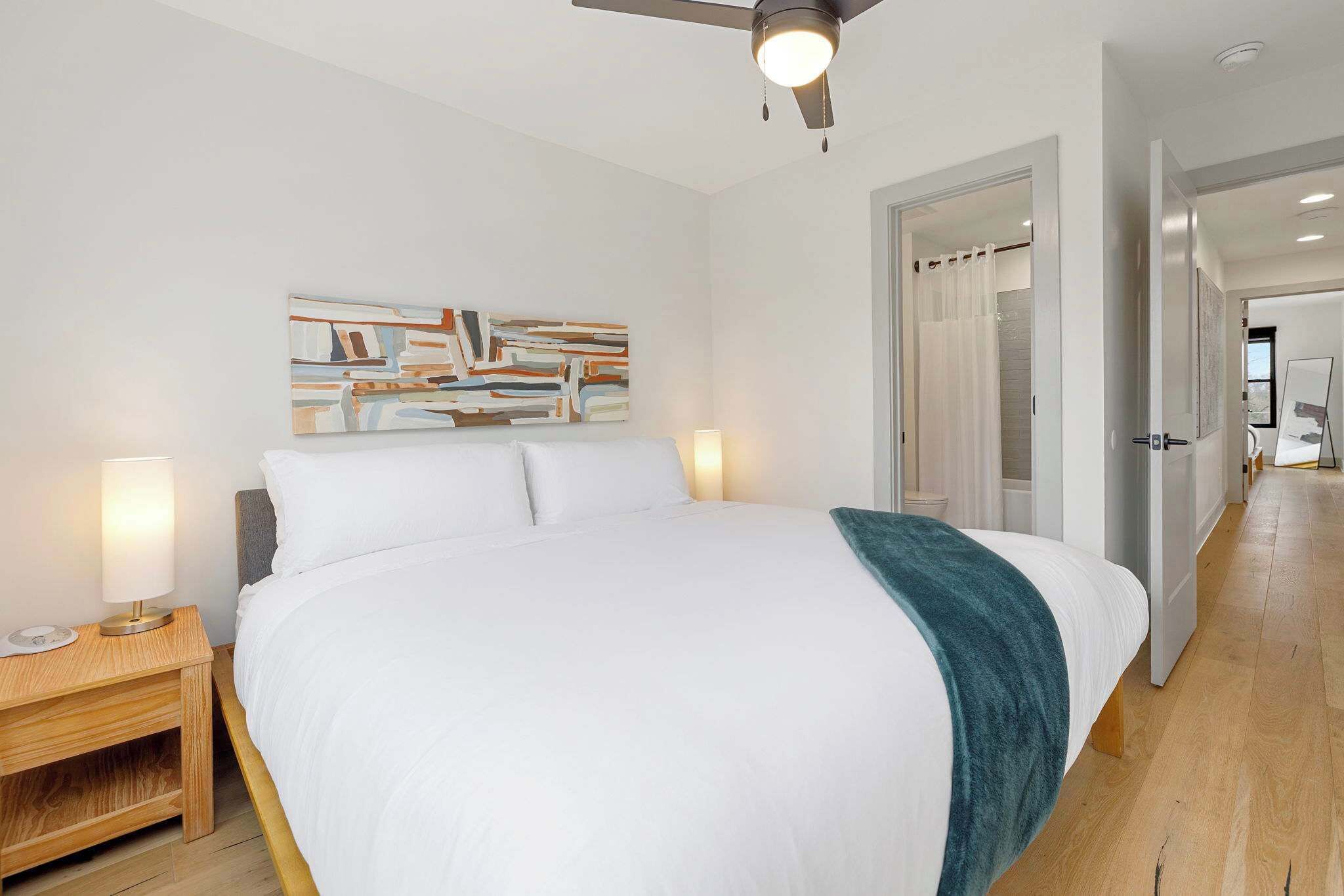 Una habitación de hotel moderna con una cama grande, una mesita de noche con lámpara, un ventilador de techo y un cuadro en la pared.