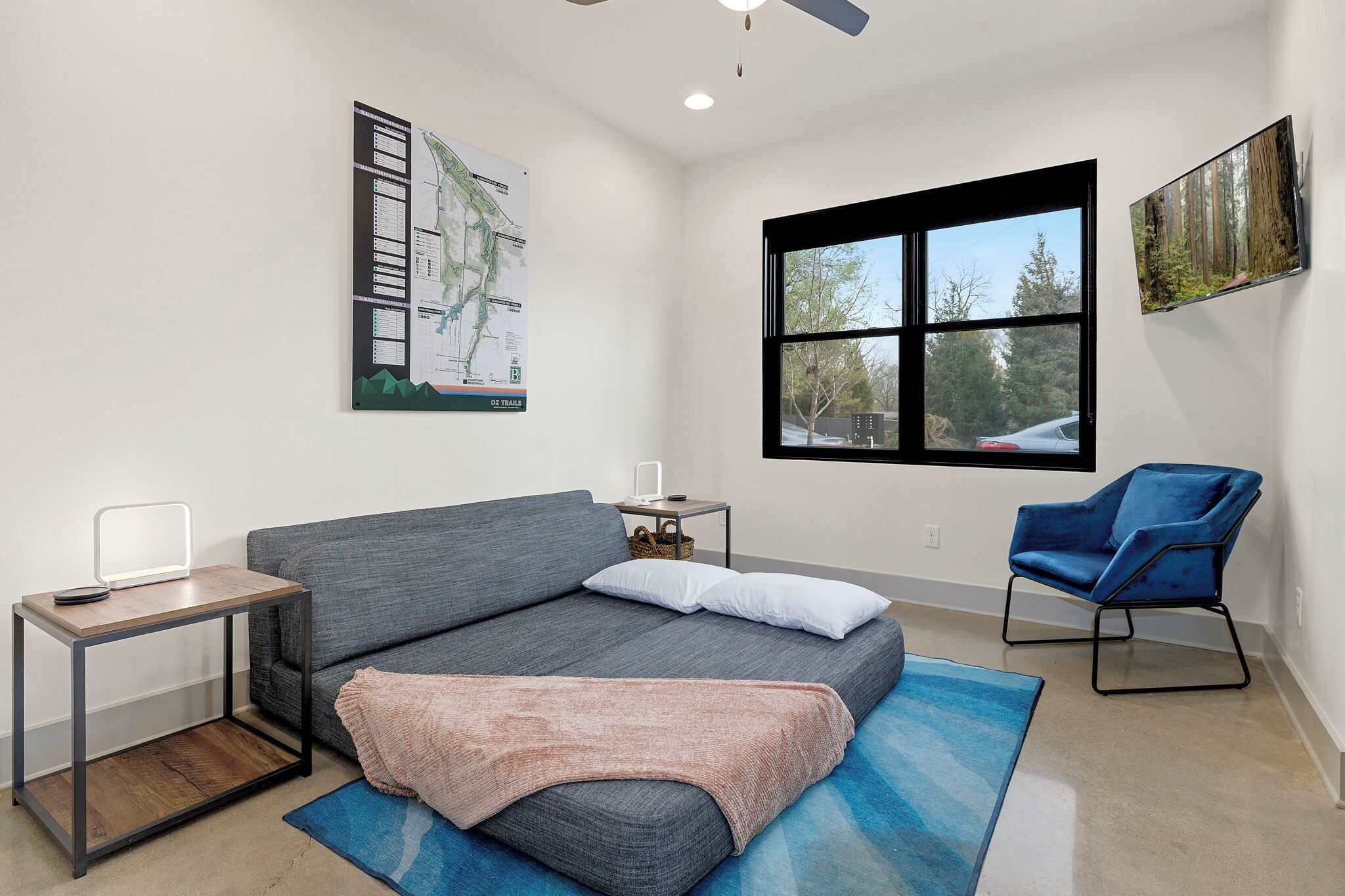 A modern room with a grey sofa, a small table with a lamp, a blue chair, a large window with a view of trees, and a map on the wall.