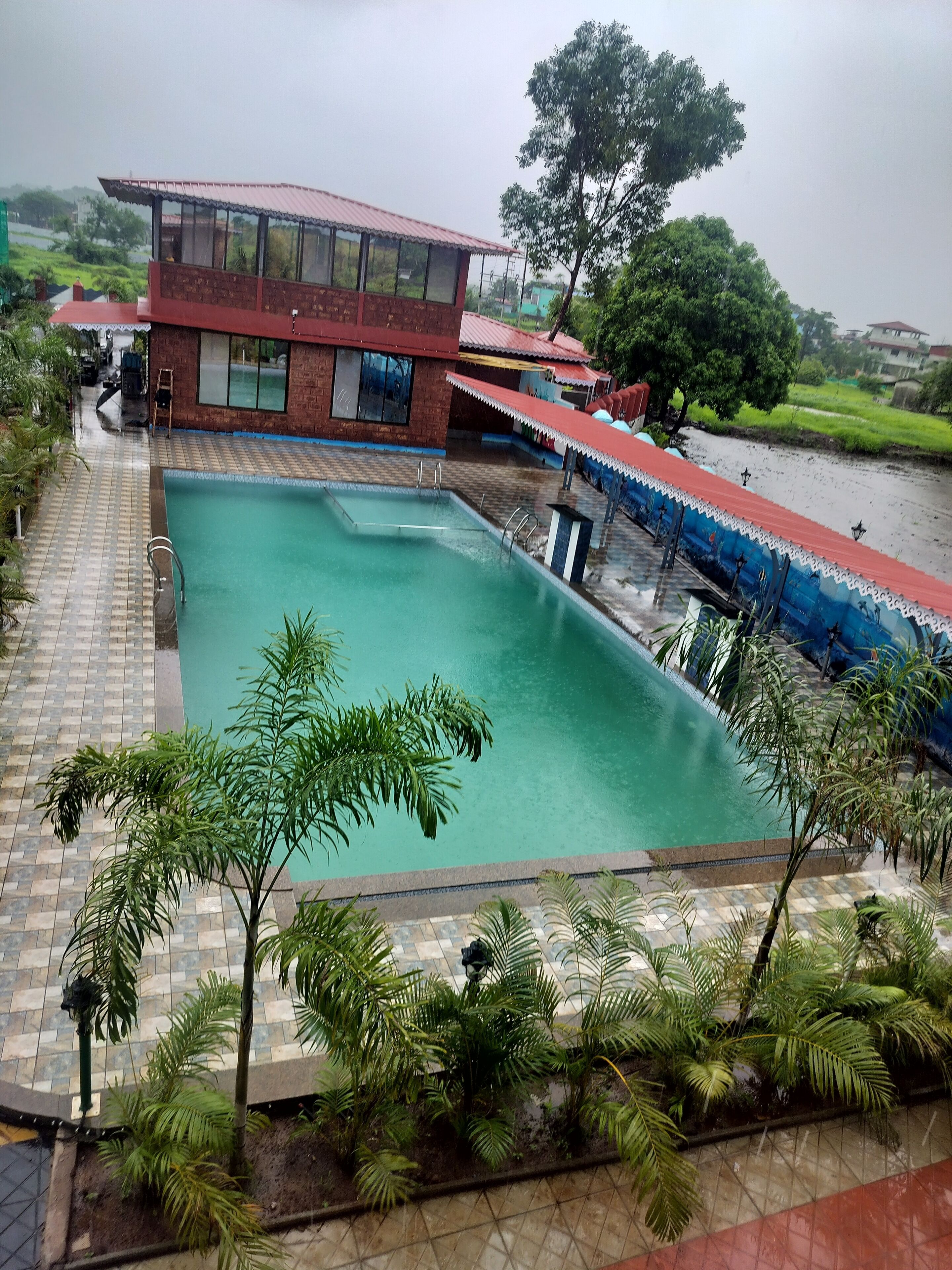 A swimming pool with a tiled deck, surrounded by palm trees and a building with a red roof.