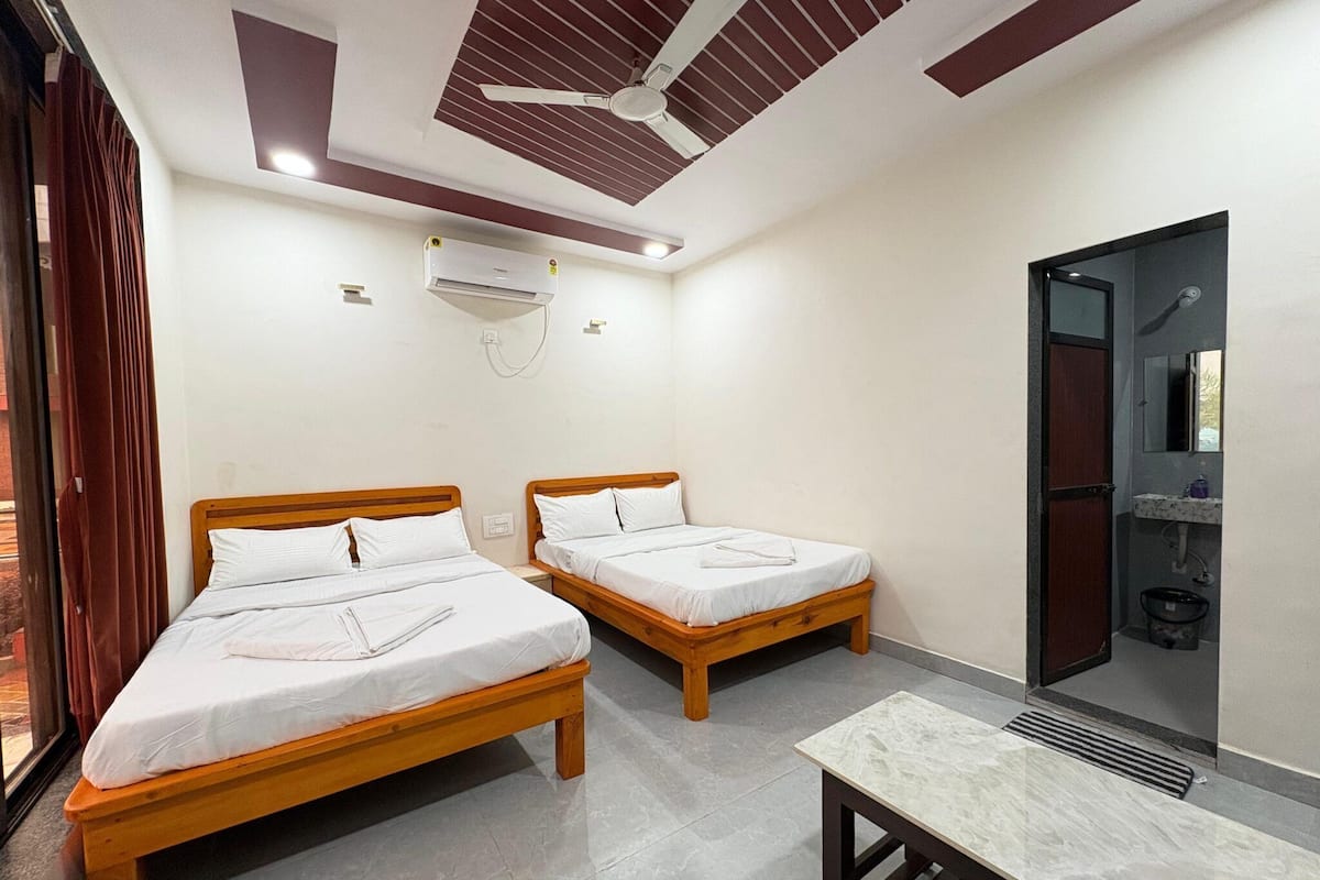 Une chambre d’hôtel avec deux lits en bois, un ventilateur de plafond, un appareil de climatisation, une petite table et une salle de bain avec un miroir et un lavabo.