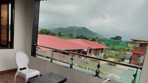 A balcony with two white plastic chairs and a dark wooden table, overlooking a view of buildings with red roofs and a green landscape. - Sandalwoods Resorts (Karjat)