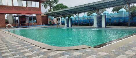 A swimming pool with a tiled deck, a covered area with a mural, and a building with large windows.