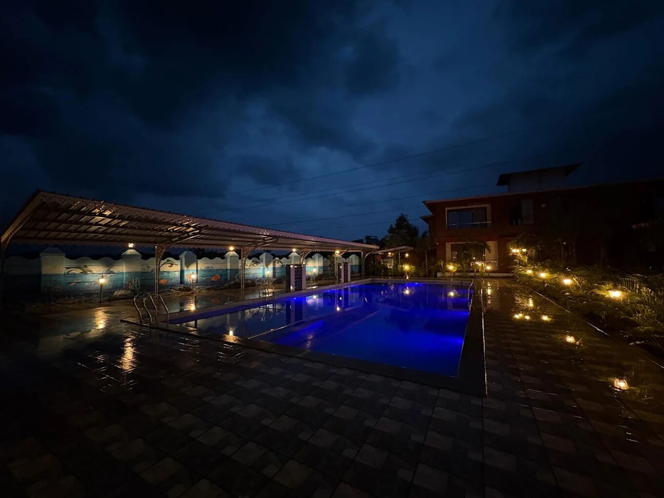 A swimming pool with a tiled deck, a covered area with seating, and a building with multiple windows.