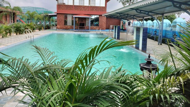 A swimming pool with a covered area, surrounded by palm trees and a building with a red roof. - Sandalwoods Resorts (Karjat)
