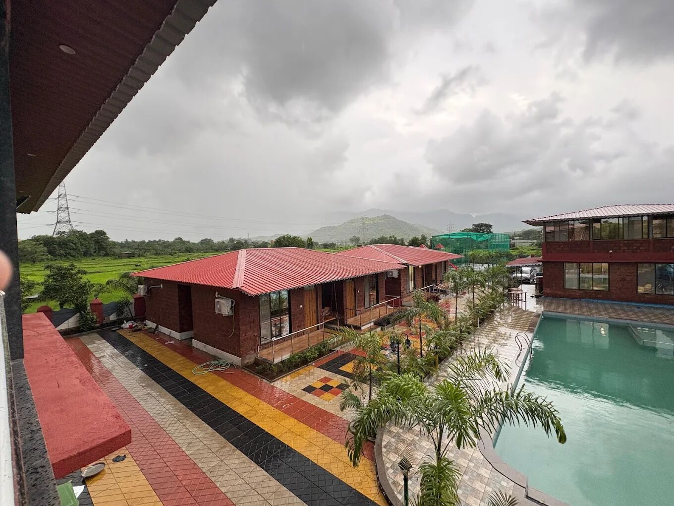 A hotel with a swimming pool, a red-roofed building, and a view of green fields and mountains.