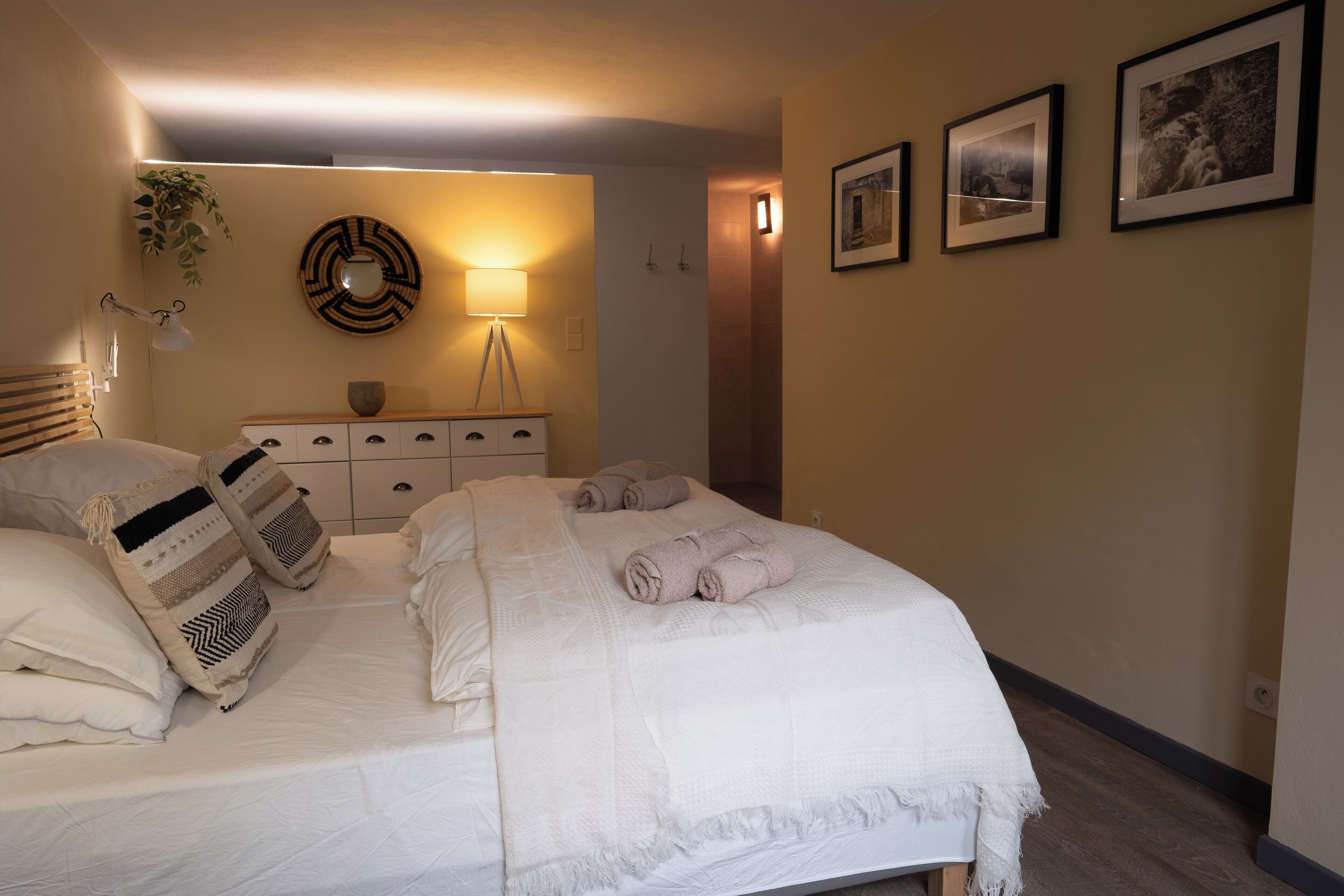 A neatly made bed with white linens and rolled towels, a bedside lamp, a wall-mounted mirror, and a small wooden dresser with a bowl on top.