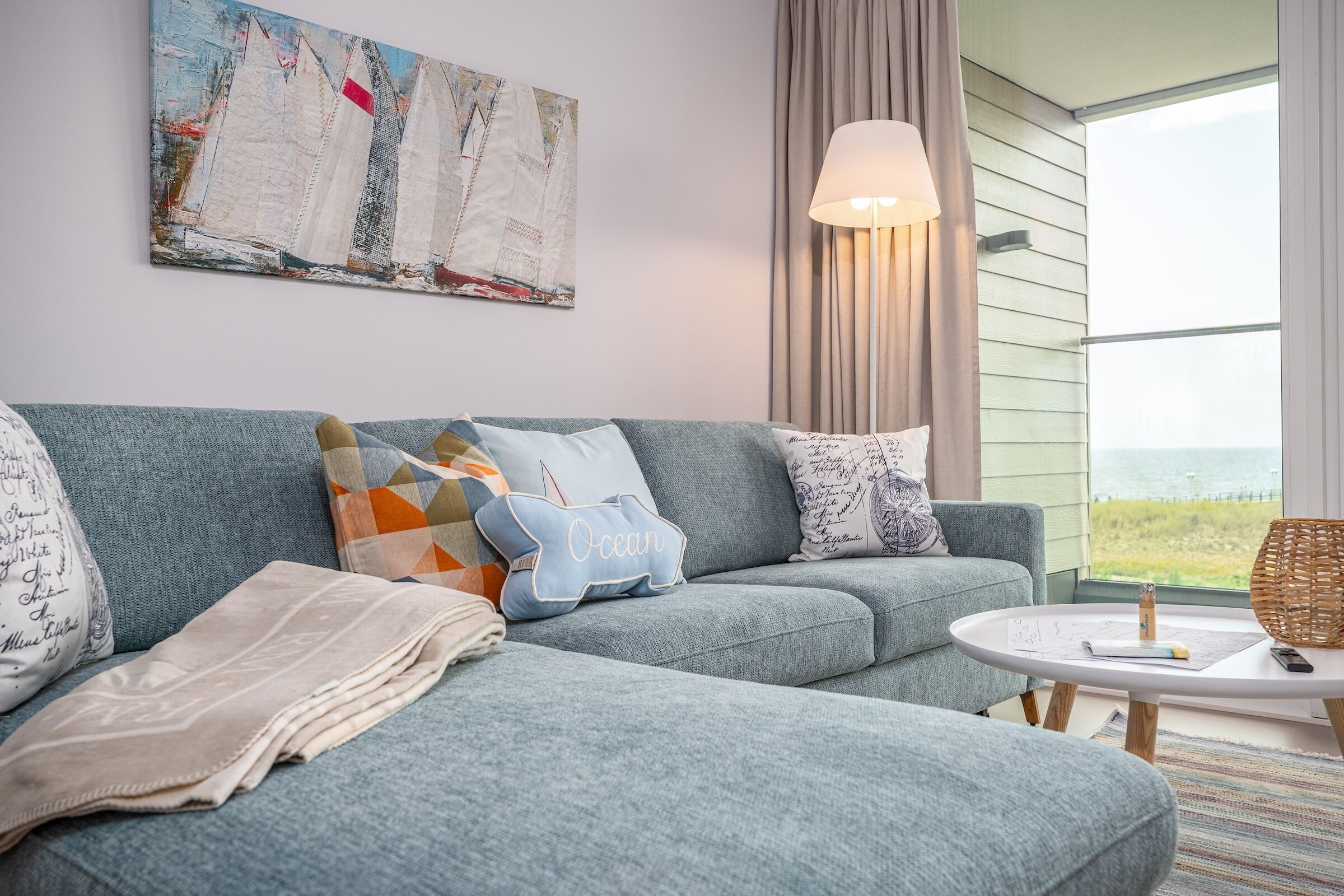 Uma sala de estar moderna com um sofá cinza, almofadas estampadas, uma mesa de centro redonda e uma janela grande com vista para o exterior.