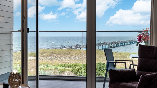 Een kamer met een grote schuifglazen deur die uitkomt op een balkon met uitzicht op zee en een pier.