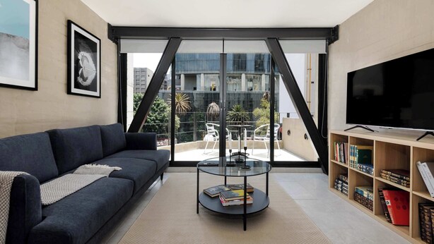 Ein modernes Wohnzimmer mit einem dunklen Sofa, einem Glastisch mit Büchern, einem Fernseher auf einer Holzablage und einem Balkon mit Gartenmöbeln.
