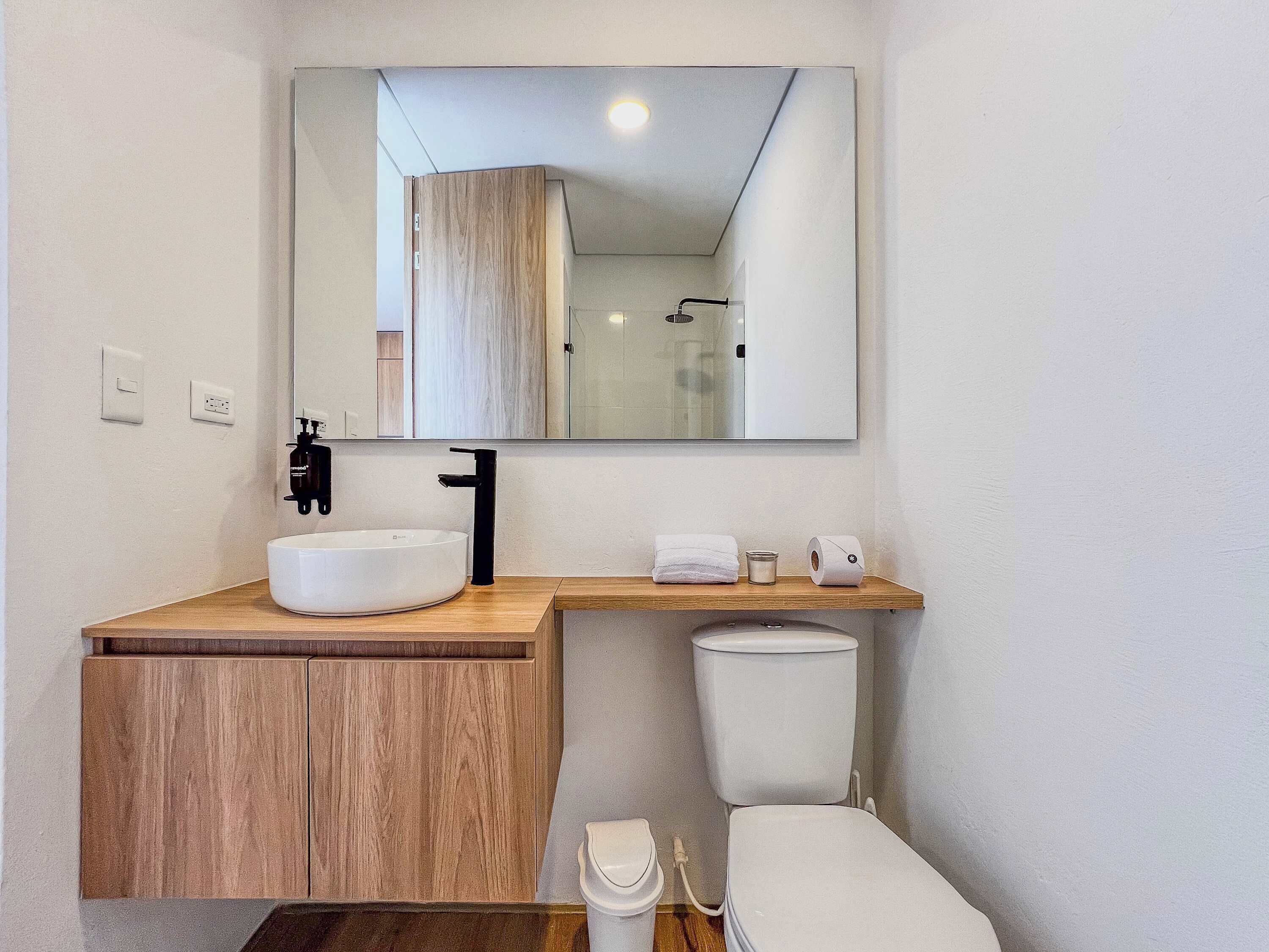 A modern bathroom with a wooden vanity, a white sink, a black faucet, a mirror above the vanity, a toilet, and a small shelf with toiletries.