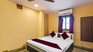 Une chambre d’hôtel avec un lit, un ventilateur de plafond, un appareil de climatisation, une fenêtre avec des rideaux violets et une armoire.
