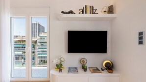 A modern living room with a large TV mounted on the wall, a white console table with decorative items, and a view of a cityscape through a glass door. - Michalakopoulou Cover & Bright Apartment (Athens)