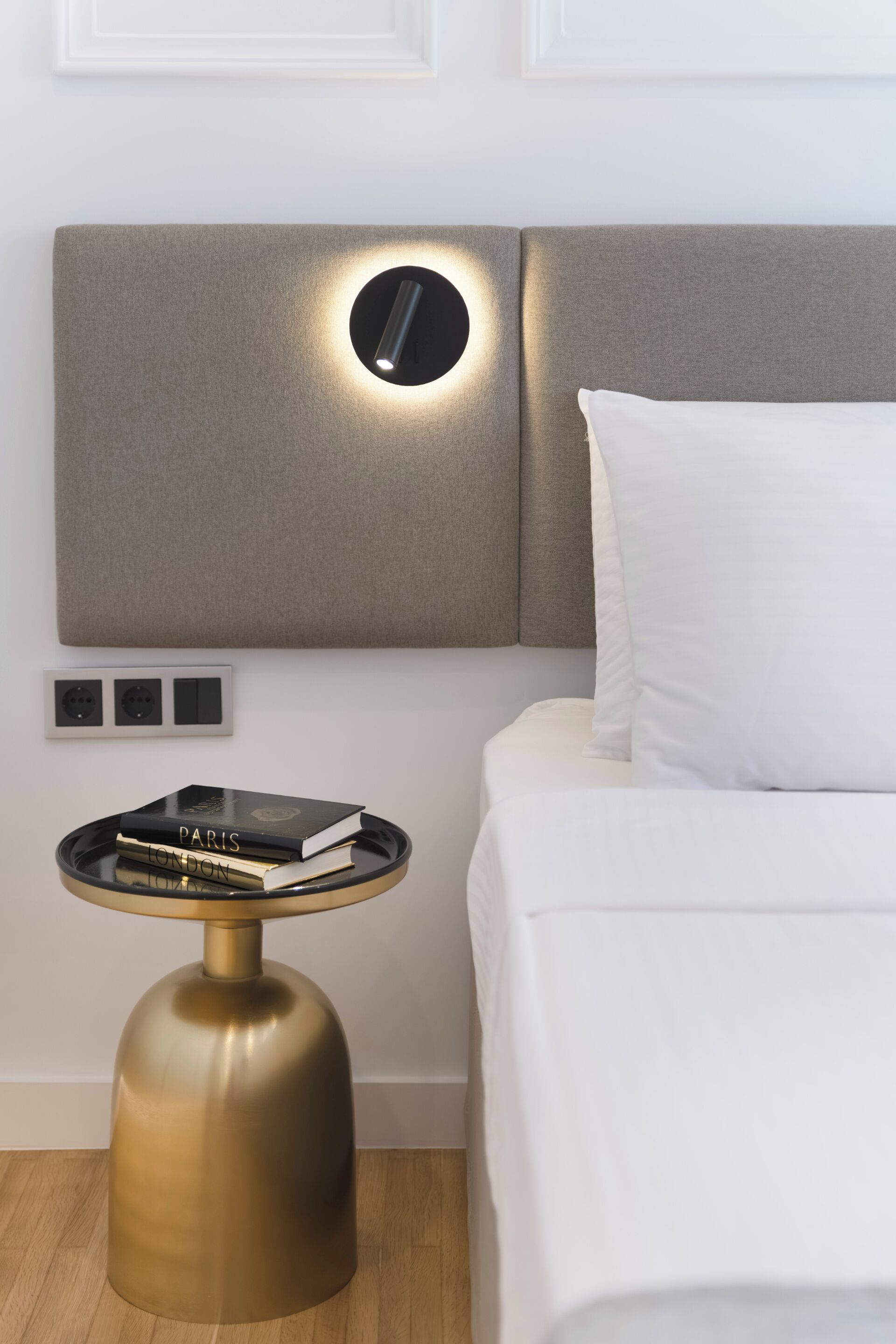 A modern hotel room with a neutral color scheme, featuring a bed with white bedding, a round gold side table with books, and a wall-mounted light fixture.