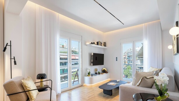 A modern living room with a sofa, armchair, and coffee table, large windows with curtains, and a mounted TV. - Michalakopoulou Cover & Bright Apartment (Athens)