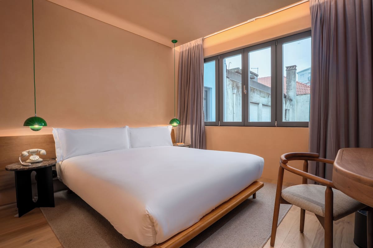 Quarto de hotel com uma cama grande, uma mesa de cabeceira com telefone, uma cadeira e uma janela com cortinas.