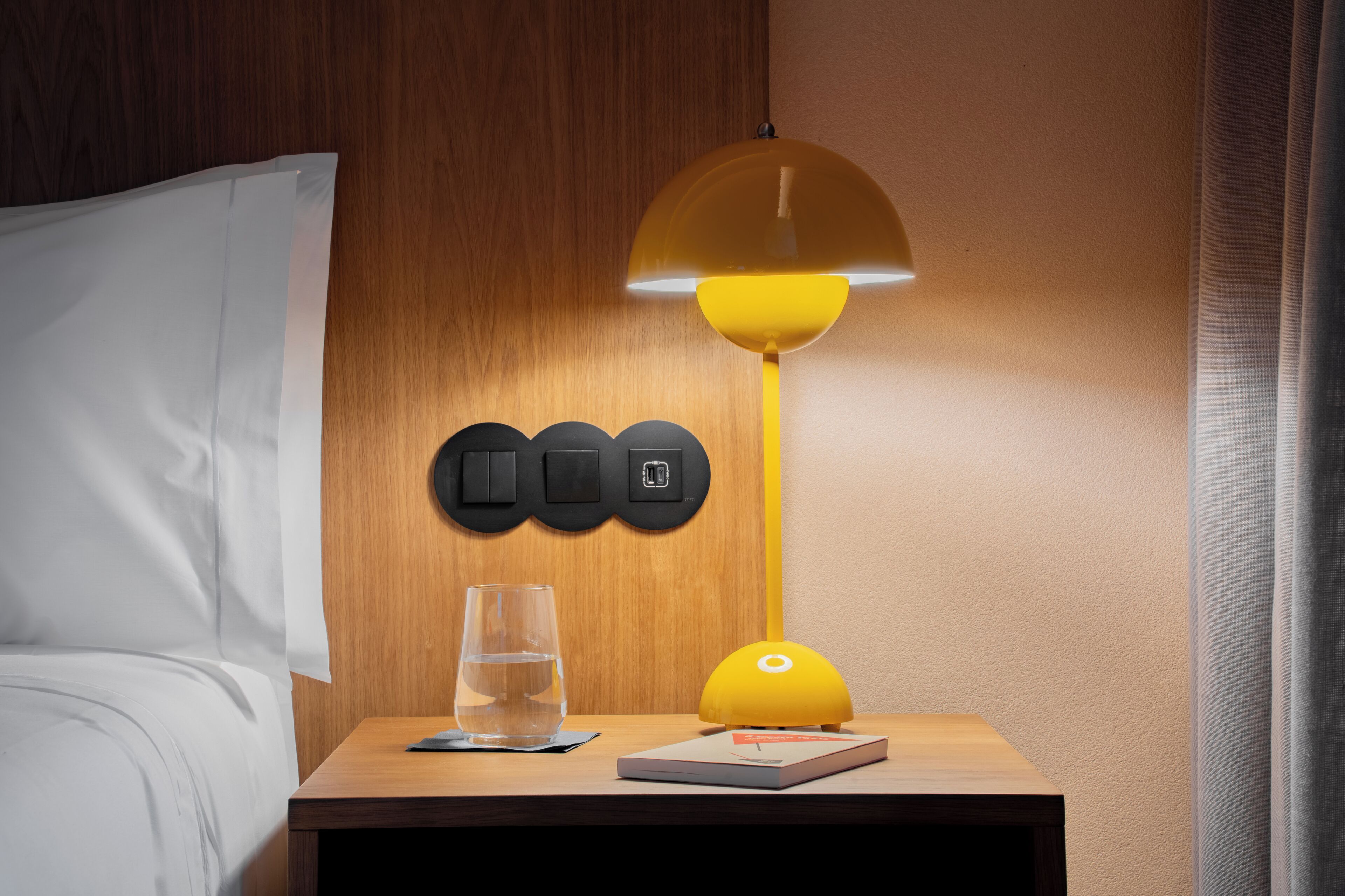 En snyggt bäddad säng med vita sängkläder, ett nattduksbord med ett glas vatten, en bok och en gul lampa med en kupformad skärm.